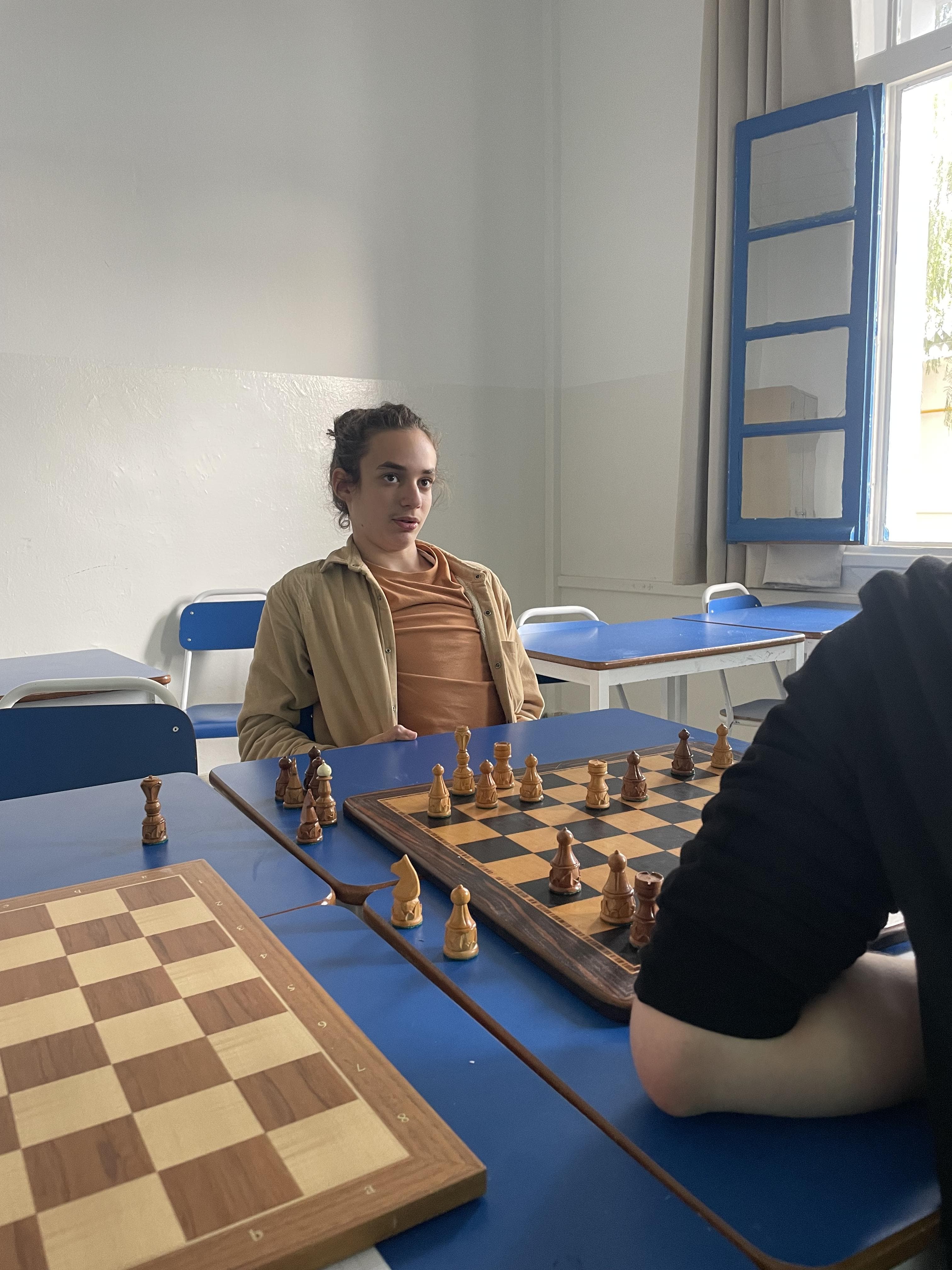 Two people seated across a chessboard in a classroom.