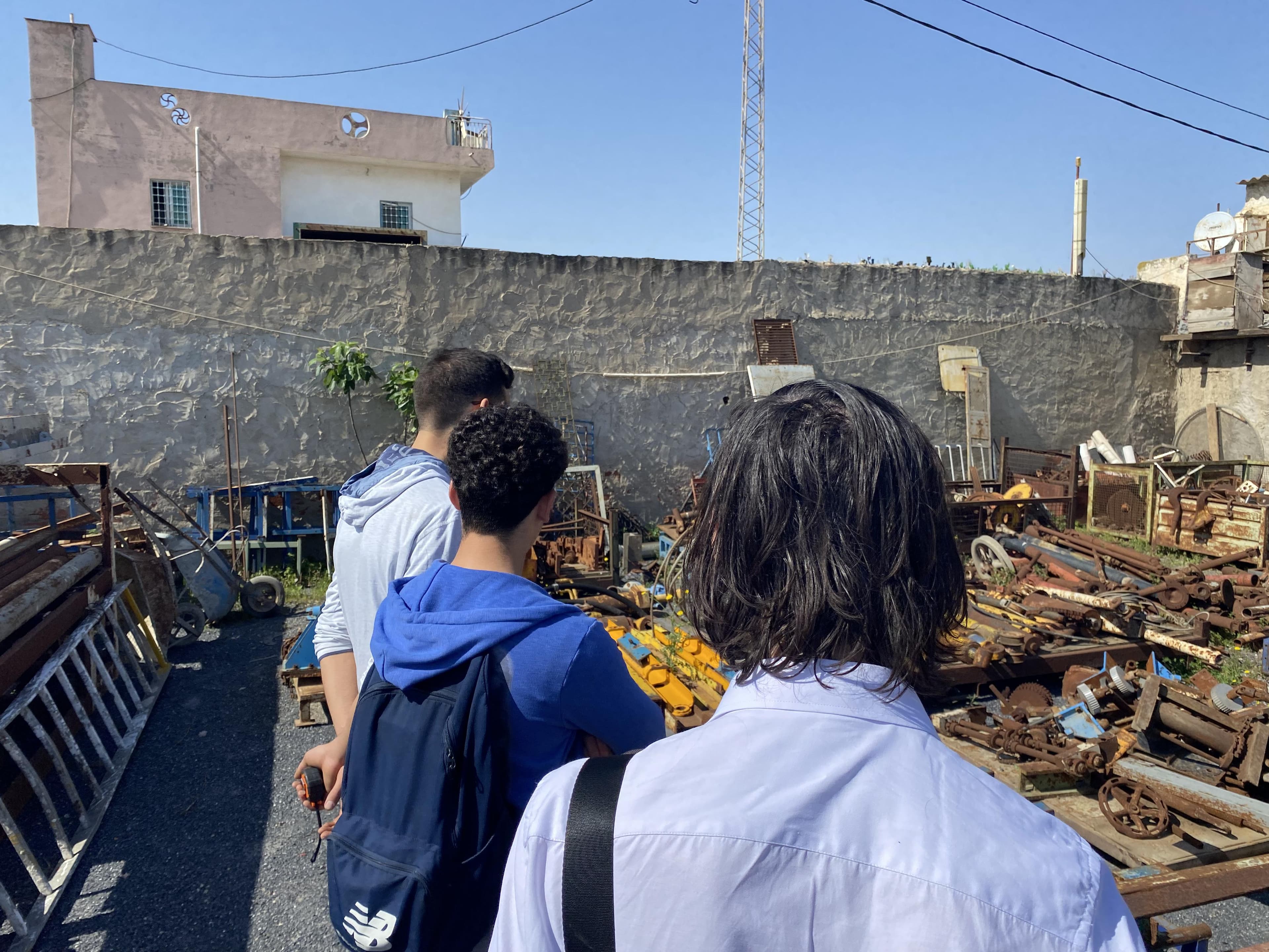 People walking through an outdoor storage area with metal parts.