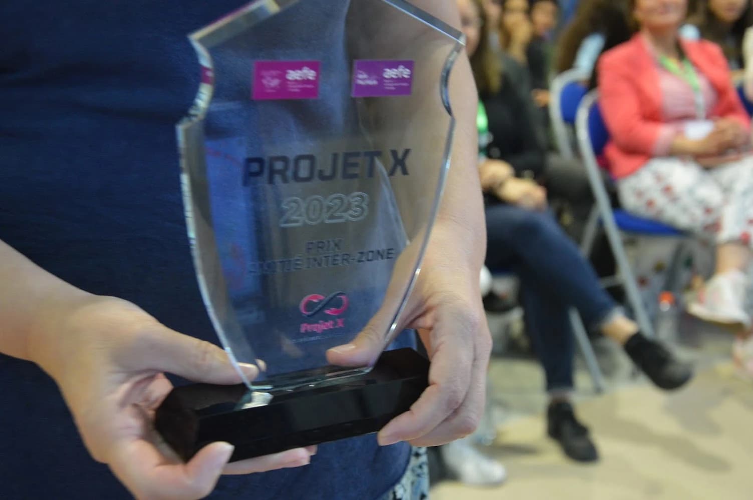 A glass trophy engraved “Projet X 2023” and “Prix lycéen inter-zone”, held in two hands