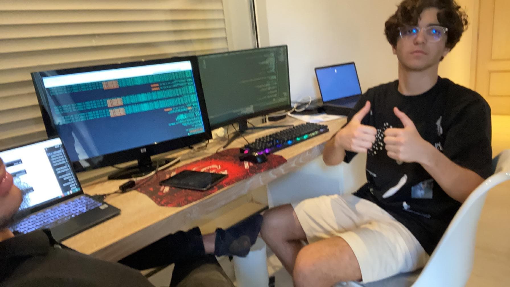 A person giving a thumbs-up beside a desk with computer screens.